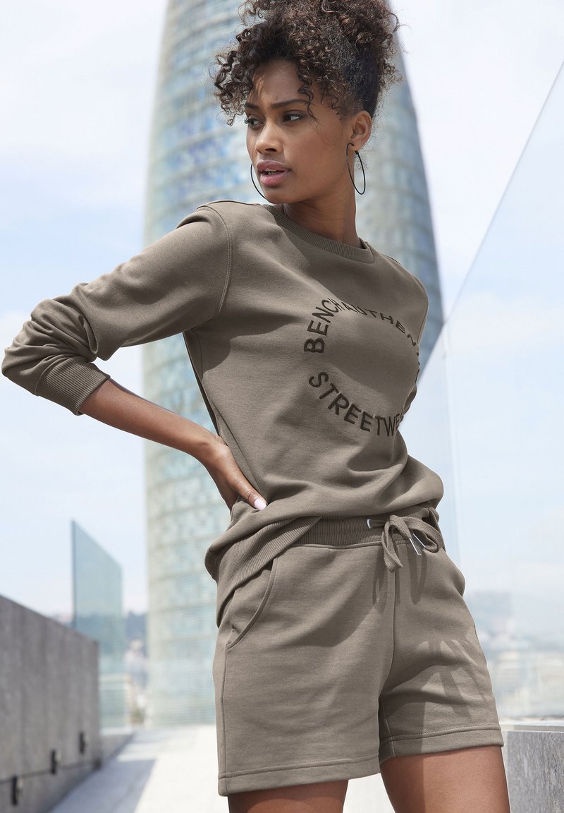 Olivgrüner Pullover und passende Shorts aus weichem Material, mit einem kreisförmigen Logo-Design auf der Vorderseite, seitlichen Taschen und einem Kordelzugbund.
