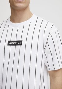 Bílá tričko s černými vertikálními pruhy, na přední straně s obdélníkovým logem "ARCHIVE" v černé barvě. Krátké rukávy, bavlněný materiál.