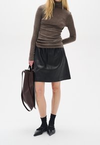 Brun tröja med hög krage, svart läderkjol, mörka strumpor och spetsiga svarta skor. Brun handbag hålls i ena handen.