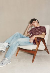 Bordová košeľa s krátkym rukávom a geometrickým vzorom, svetlomodré džínsy a sivé tenisky. Drevená stolička s béžovým čalúneným sedákom.