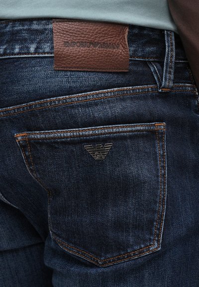 Temno modri jeans iz denima imajo usnjen logotip v rjavi barvi na pasu in kovinski emblem na zadnjem žepu z oranžnimi šivi.