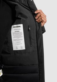 Black padded jacket interior with a zippered pocket, care label, and fabric texture visible. Features a sleek design and functional hardware.