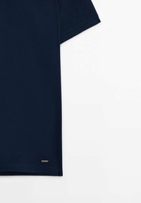 Chemise bleu foncé à manches courtes avec une petite étiquette métallique rectangulaire de marque près de l'ourlet inférieur sur fond blanc.