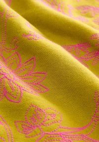 Tissu brodé avec un fond jaune vif et des motifs floraux roses élaborés, présentant une texture lisse et douce.