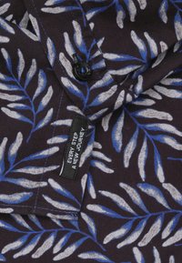 Korte mouwen shirt in donker stof met blauwe en witte bladdessins, zwarte knop, en een label met de tekst "IEDERE STAP EEN NIEUWE REIS."