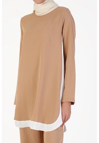 Tunique beige à manches longues avec un ourlet incurvé et une accentuation blanche en bas. Réalisée en tissu lisse, elle présente un col rond simple et une coupe décontractée.