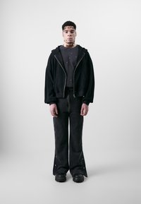 Sort hættejakke med lynlås og brede ærmer over en sort T-shirt, parret med buksflare sorte bukser med trykknapsdetaljer og struktureret stof.