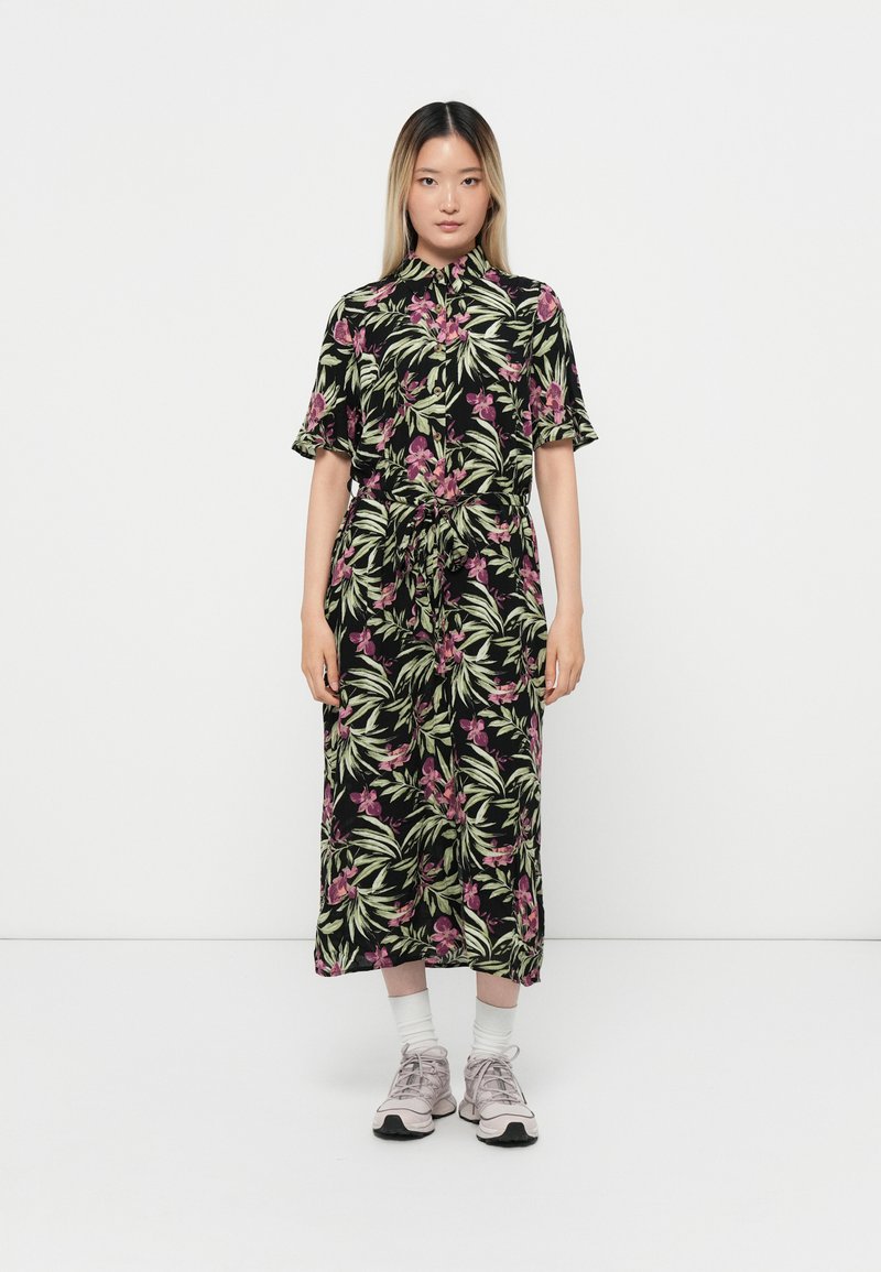 Robe noire à motifs floraux avec des orchidees roses et des feuilles vertes, dotée d'une taille ceinturée et de manches courtes, associée à des baskets légères roses.