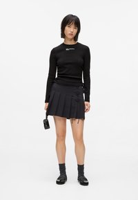 Czarny top z długim rękawem z logo, w zestawieniu z czarną plisowaną mini spódnicą z lace-up. Model ma na sobie czarne buty i szare skarpetki.