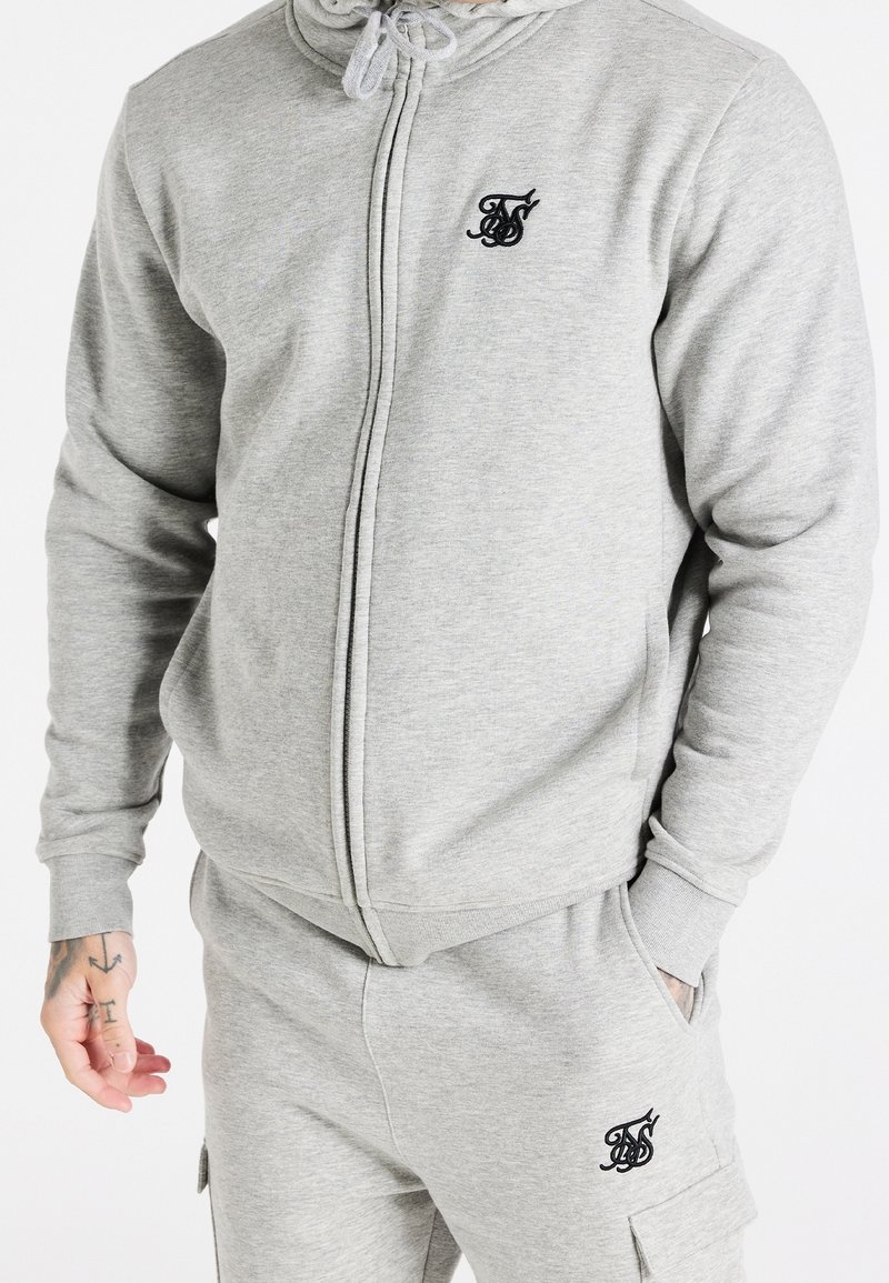 Felpa grigia con zip realizzata in tessuto morbido, caratterizzata da un piccolo logo nero sul petto, con cappuccio regolabile e chiusura a zip.