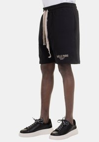 Shorts neri con vita elasticizzata e cordoncino beige, con la scritta "GÄELLE PARIS" stampata in oro. Abbinati a scarpe da ginnastica nere con suola bianca.