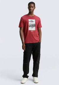 Sarkana kokvilnas T-krekls ar kalnu grafiku, apaļu kakla izgriezumu un īsām piedurknēm, kombinēts ar vaļīgām melnām cargo biksēm un baltām kedām.