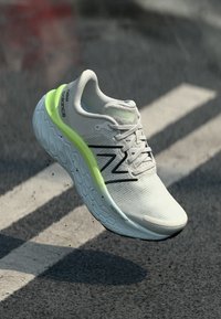 Light gray New Balance running shoe with neon green accent and textured sole floating above asphalt with white road markings.