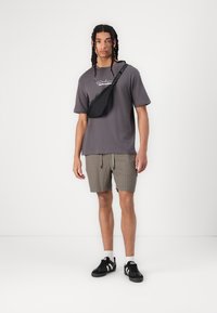 Gray short-sleeve t-shirt with white logo, tan plaid shorts, black crossbody bag, and black sneakers with white accents.
