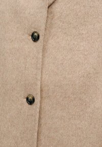 Ανοιχτό καφέ παλτό από μείγμα μαλλιού με απαλό υφή, που διαθέτει δύο μαύρα κουμπιά και γιακά με λοξή εγκοπή.