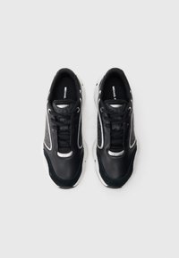 Une paire de baskets Michael Kors noires et grises avec des semelles blanches, des lacets noirs et des semelles intérieures de marque sur un fond blanc.