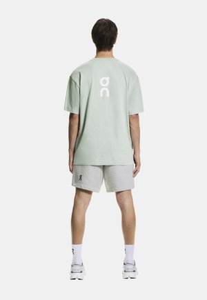 Mann trägt hellgrünes übergroßes T-Shirt und graue Shorts mit Logos, weiße Socken und Sneakers, steht mit dem Rücken zum Betrachter vor einem einfarbigen Hintergrund.