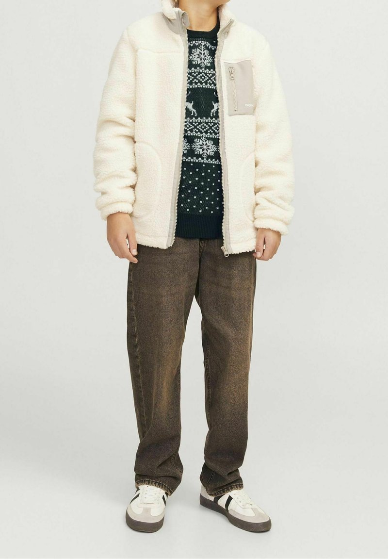 Personne portant une veste polaire blanche par-dessus un pull sombre à motifs, un pantalon marron ample et des baskets blanches avec des bandes noires.