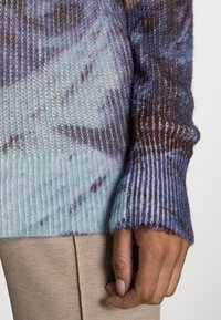 Main dans le repos sur le côté, portant un chandail en tricot doux à manches longues avec des poignets côtelés dans des tons de bleu et de violet sur un pantalon beigne.