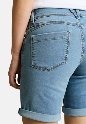 Vista trasera de una persona que lleva pantalones cortos vaqueros azul claro con los bajos doblados y una camiseta blanca, mano apoyada al lado del muslo.