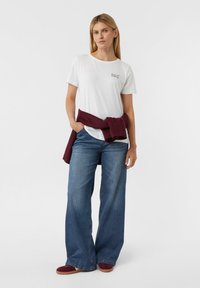 Weißes Baumwoll-T-Shirt mit einem kleinen Aufdruck, kombiniert mit weiten blauen Denim-Jeans. Ein kastanienbrauner Pullover ist um die Taille gebunden. Rote Wildlederschuhe.
