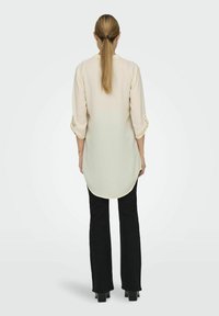 Light yellow long-sleeve shirt with buttoned cuffs, featuring a rounded hem. Paired with black flared pants and glossy footwear.