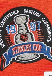 Toppa della Stanley Cup datato 1997 con il logo NHL, il testo "Western Conference" e "Eastern Conference" su un tessuto arancione.