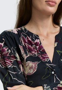 Kvinna som bär en mörkblommig blus med vinröda och beige blommor, synlig hals, läppar och korslagda armar.