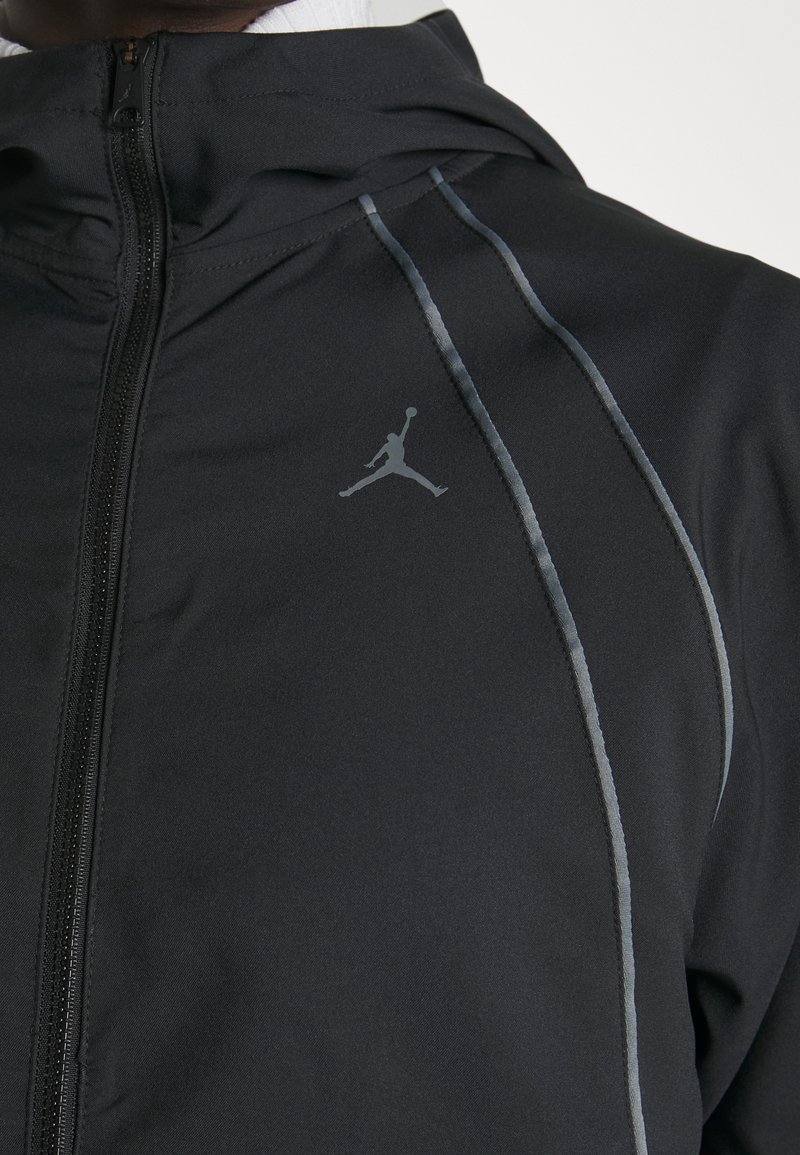 Chaqueta negra con cremallera y piping reflectante, además de un logo gris de Jumpman en la parte superior del pecho, llevada por una persona.
