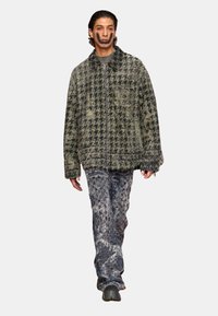 Veste à motif pied-de-poule vert avec une fermeture éclair, associée à un jean texturé bleu-gris. Le mannequin porte des chaussures noires.