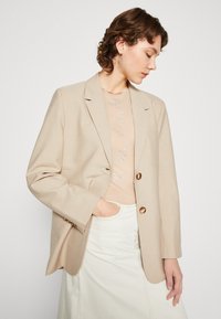 Μπεζ σακάκι με δύο χρυσά κουμπιά, δομημένος σχεδιασμός ώμου και λεπτή υφή. Φορεμένο πάνω από ένα ανοιχτό μπλουζάκι με διακοσμητικό ασημένιο μοτίβο.