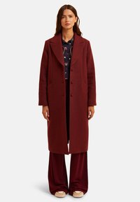Manteau long en laine bordeaux avec un col, deux poches frontales, trois boutons et une coupe droite. Porté sur une chemise à motifs et un pantalon ample.