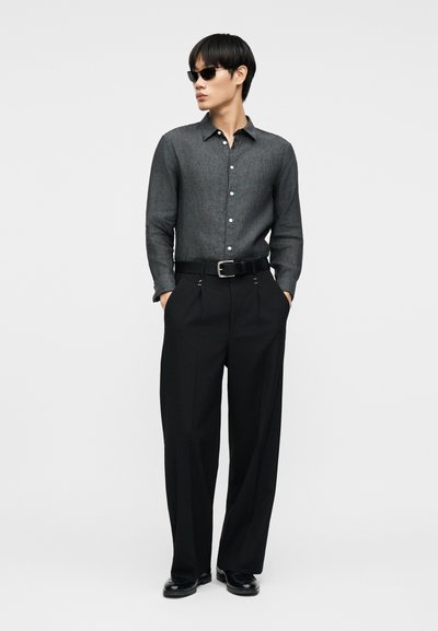 Άνδρας που φοράει σκούρο γκρι πουκάμισο με κουμπιά, μαύρο παντελόνι φαρδύ, μαύρη ζώνη, μαύρα παπούτσια και γυαλιά ηλίου, στέκεται με τα χέρια στις τσέπες.
