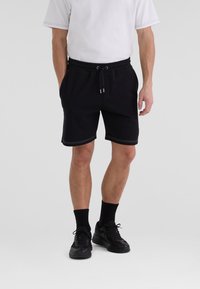 Svarta shorts i mjuk tyg med elastisk midja, med vita sömmar som detaljer. Bärs med svarta sneakers och ankelsockor.