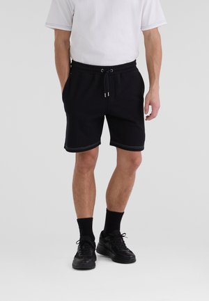 Svarta shorts i mjuk tyg med elastisk midja, med vita sömmar som detaljer. Bärs med svarta sneakers och ankelsockor.
