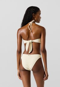 Žena s dlouhými kudrnatými vlasy, oblečená v krémovém bikinách s texturou uvázaných vzadu, stojící na jednoduchém světle šedém pozadí.