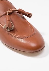 Bruine lederen schoen met een ronde neus, decoratieve franjes en stiksels langs de rand, met een gladde, licht glanzende textuur.
