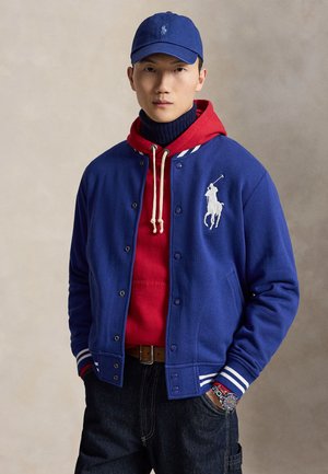 Homme portant une casquette bleue, une veste bleue avec un logo blanc, un sweat à capuche rouge, un jean foncé et une montre-bracelet, debout les mains dans les poches devant un fond neutre.