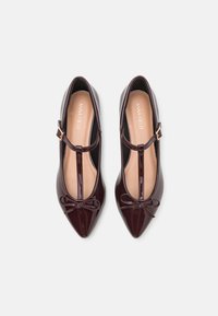 Chaussures en cuir verni bourgogne avec un bout pointu, une sangle en T, un accent en nœud à l'avant et une semelle intérieure nude légèrement rembourrée.