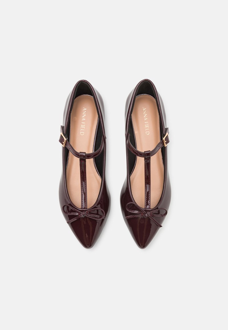 Scarpe in pelle verniciata bordeaux con punta affusolata, cinturino T, dettaglio a fiocco nella parte anteriore e soletta nuda leggermente imbottita.