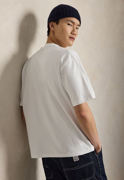 Fiatal férfi fehér pólóban és fekete kötött sapkában, enyhén fordulva, kezével a zsebekben, egyszínű bézs háttér előtt.