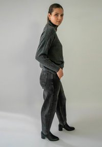 Jeune femme en pull à col roulé gris, pantalon noir taille haute et bottes à talons noires, se tenant de côté contre un fond clair uni.