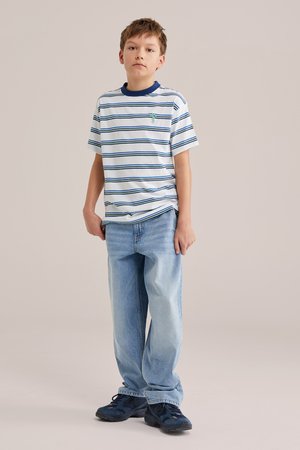 Niño de pie con camiseta blanca con rayas azules, jeans azul claro y zapatillas oscuras, frente a un fondo liso.