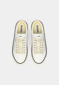 Une paire de baskets blanches basses avec des lacets beiges, des bordures noires et le logo "HUGO" sur les semelles intérieures et les côtés, présentée de dessus sur un fond blanc.