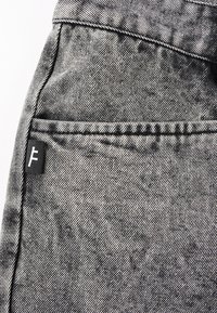 Svart og grå denimstoff med en synlig lomme og stikninger. Har et lite svart merke med en hvit logo. Teksturert overflate.