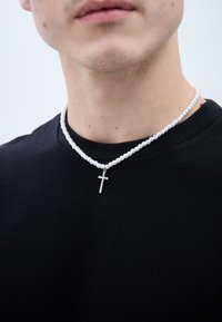 Pērļu kaklarota ar sudraba krustu piekariņu, kas valkāta uz melna T-krekla. Kaklarotā ir baltas, apaļas pērles un spīdīgs, metalisks apdare.