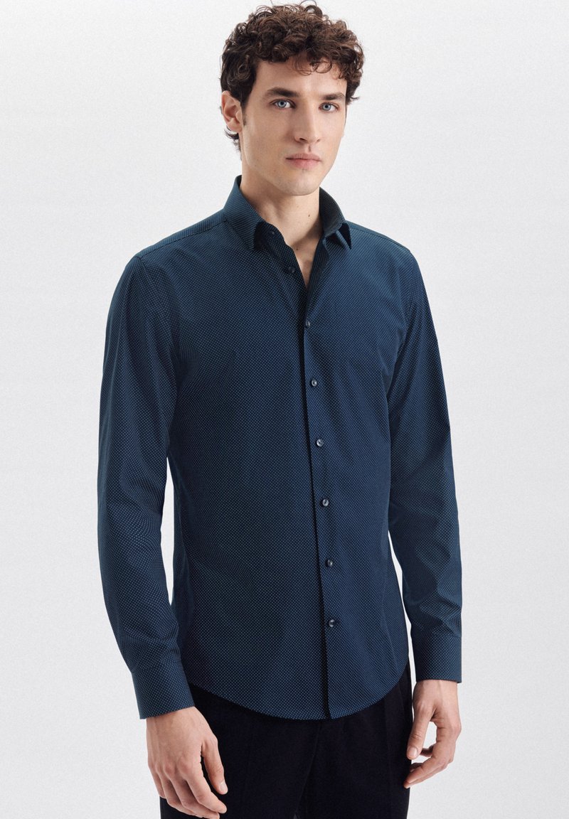 Mørkeblå langærmet skjorte med et subtilt prikkemønster, knaplukning, buet bund og knapmansjetter. Glat stof med en let glans.