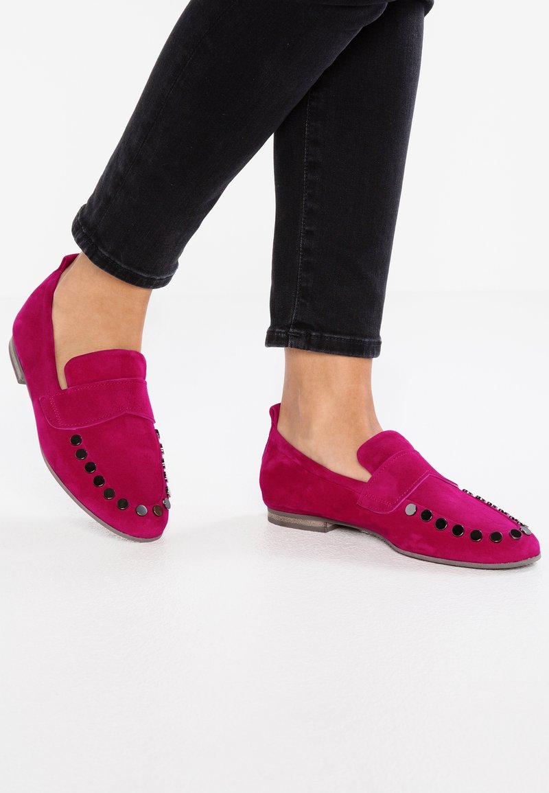 Fødder iført klare pink ruskind loafers med sorte nitter langs tæerne, parret med sorte cropped bukser, på et hvidt gulv.