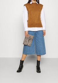 Pull sans manches marron par-dessus une chemise blanche à col, associé à des culottes en denim. Des bottines noires et une pochette imprimée léopard complètent le look.