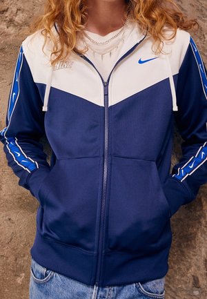 Osoba nosząca granatowo-białą kurtkę Nike zapinaną na zamek błyskawiczny z niebiesko-białymi wzorzystymi rękawami oraz jasnymi jeansami na tle szorstkiej ściany.